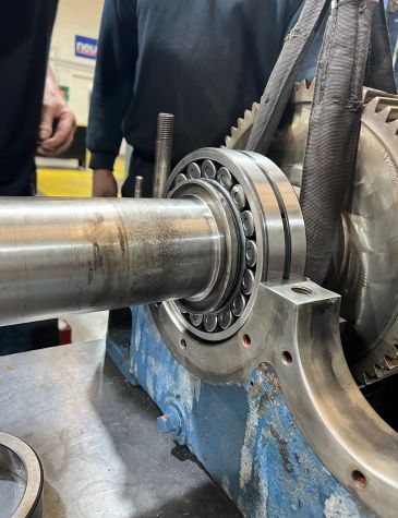 A close-up shot of an industrial motor and gearbox assembly. A new roller bearing has been installed onto the shaft, which is ready to be seated in the gearbox casing. The assembly is resting on a workbench.