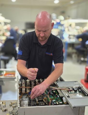 A Neutronic Technologies engineer, wearing a black polo shirt, is intently focused on repairing an electronic unit. He is using a screwdriver to work on the internal wiring and components, including circuit boards and relays.