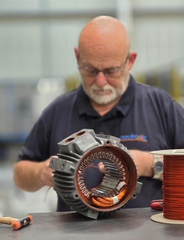 Person in a dark shirt working on an electric motor stator with visible copper windings, alongside a spool of copper wire and tools like a hammer on the workbench.