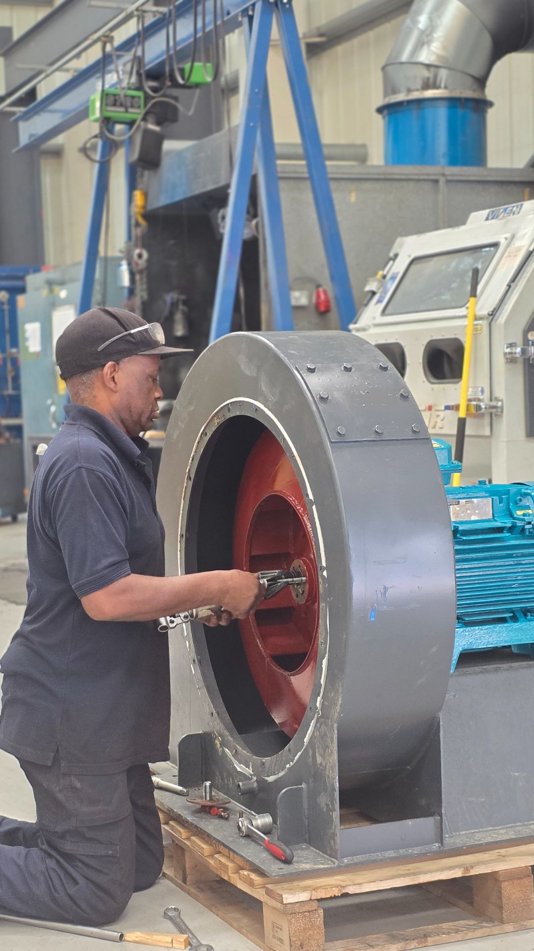 Industrial fan repair in a workshop setting, showing a technician working on a large grey and red fan housing mounted on a pallet, with tools and machinery in the background – professional industrial fan maintenance and repair services.