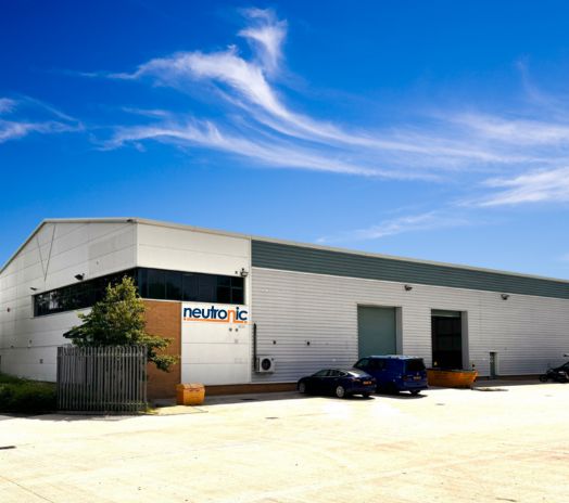 large industrial building with a white and gray exterior, labeled "neutronic" in blue and orange letters. The building has a flat roof with a slight slope, several large garage doors, and small windows. There are two cars parked in front of the building on a spacious concrete lot. The sky above is clear with some wispy clouds. Contact Neutronic Technologies for Electro Mechanical Repair.
