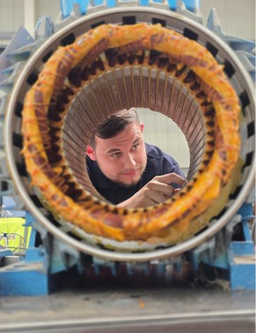 Technician inspecting the interior of a large industrial machine part with copper windings, viewed through the circular opening of a stator mounted on a blue frame