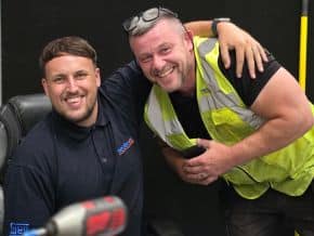 Two Neutronic team members posing in a workshop setting, one seated in a navy polo shirt with company logo, the other standing in a high-visibility vest, with tools visible in the foreground