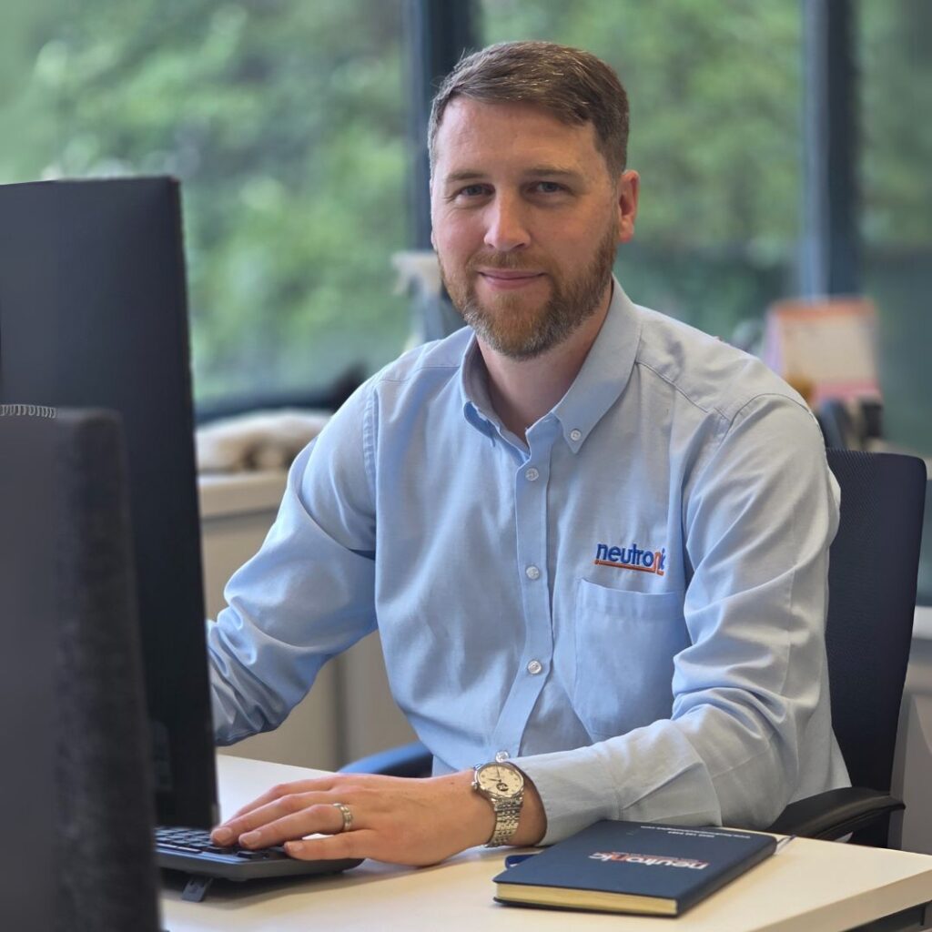 Stephen James, General Manager of Neutronic Technologies Ltd., seated at his office desk wearing a light blue branded shirt. He is working on a computer, with a watch on his left wrist. The office features large windows with greenery visible outside.