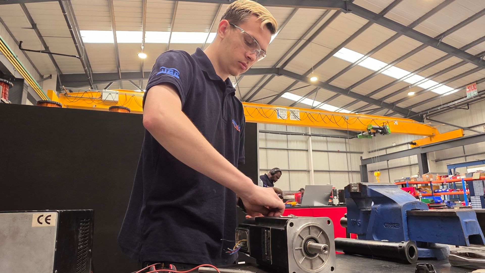 Technician conducting servo motor repair and maintenance in an industrial workshop, surrounded by tools, equipment, and supply components including a blue vise and overhead crane system.