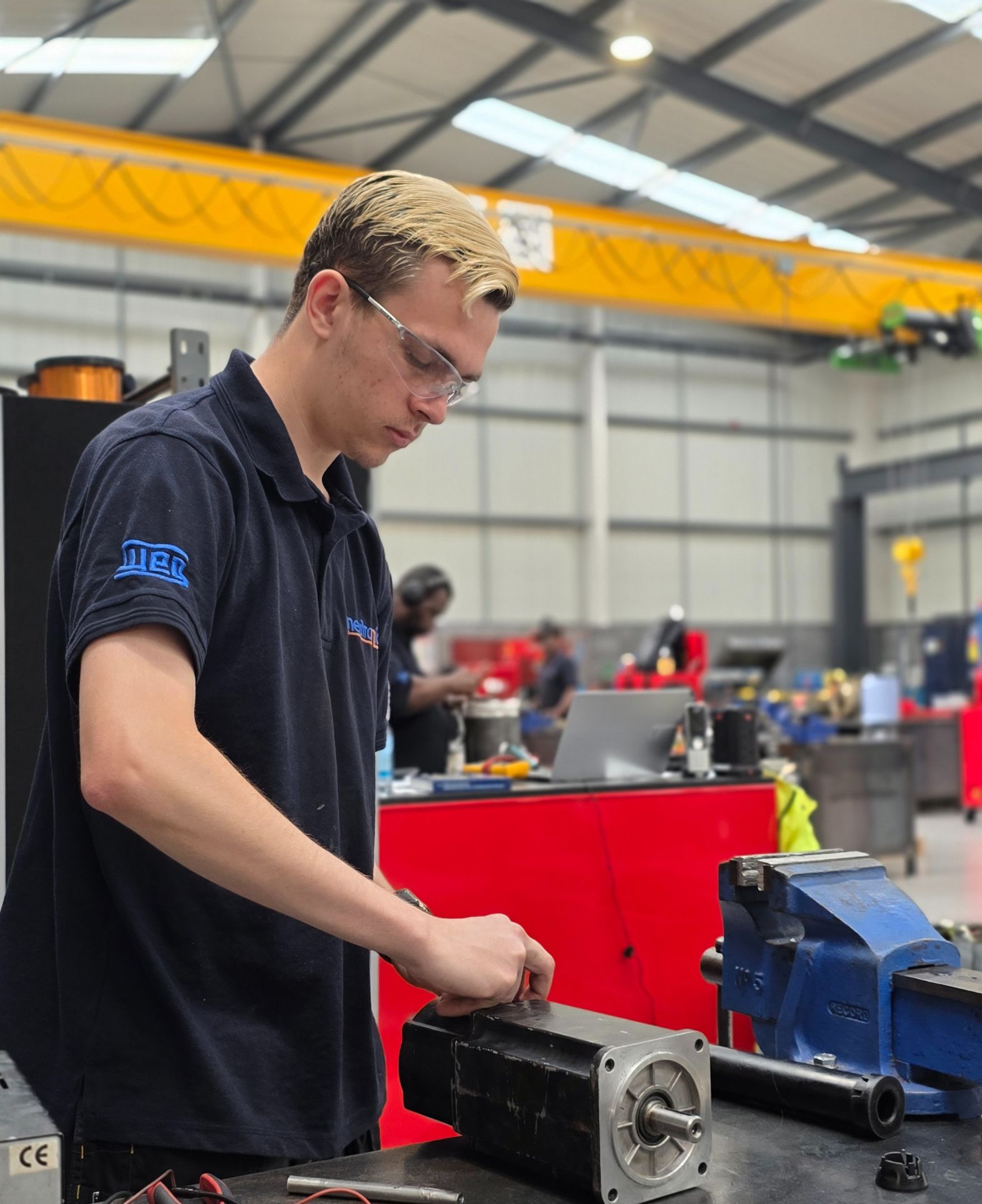 A technician performing servo motor repairs in a workshop, wearing a dark blue Neutronic Technologies uniform. The workspace includes tools, a blue vice, and an overhead crane system, emphasising a professional repair environment. Industrial servo motor repair and Servo Motor Repairs with detailed servo motor components used in servo repair services
